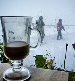 Foto de um chocolate quente e, atrás, janela embaçada com vista para o gelo.