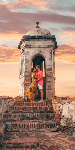 Foto de um casal se beijando em um forte antigo no pôr do sol.