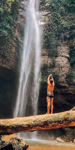 Foto de um homem com roupas de banho com as mãos para cima virado para uma grande cachoeira.