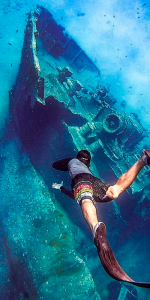 Foto submersa de um homem mergulhando com um navio naufragado no fundo do mar.