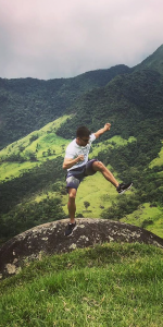 Foto de um homem pulando em cima de uma pedra com montanhas com muita grama e árvores.