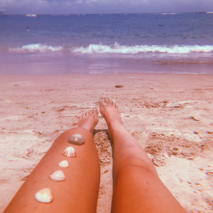 Foto das pernas de uma mulher deitada na areia da praia de frente para o mar com conchas em cima da perna esquerda