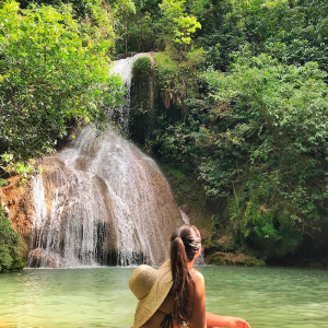 Em primeiro plano há uma menina co um chapéu pendurado nas costas olhando para a cachoeira que está ao fundo com muitas árvores no entorno.