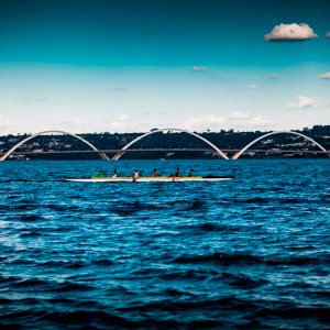 Foto do Lago Paranoá com uma ponte ao fundo e uma canoa com algumas pessoas remando dentro a luz do dia.