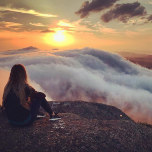 Menina de costas para a foto em cima de um pico de uma montanha em frente a um pôr-do-sol