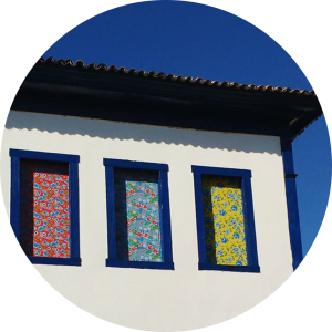 Casarão antigo, com três janelas coloridas (primeira janela com uma cortina vermelha com azul, a segunda com uma cortina azul e rosa e a terceira com uma cortina amarela e verde e paredes brancas e marcos azuis.