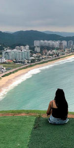 Menina sentada na ponta do morro admirando a vista que consiste em a cidade e a beira da praia com ondas.