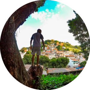 Foto de homem em cicma de pedra de frente para uma montanha com vasta vegetação e casas da região