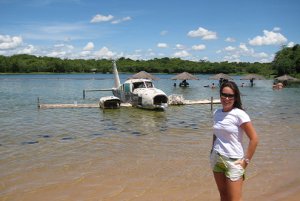 Natalia em frente a um lago com um avião antigo e castigado por uma possível queda e tempo