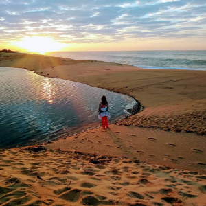 Menina em meio a areia de costas para a foto e de frente para o lago com o pôr do sol ao fundo.