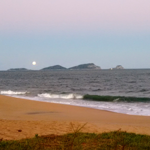 Foto da beira da praia com areia e ondas ao fundo, ao final da tarde.