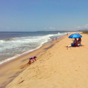 Foto da areia da praia com as ondas do mar á esquerda e uma família sentada na areia com um guarda-sol e crianças brincando.