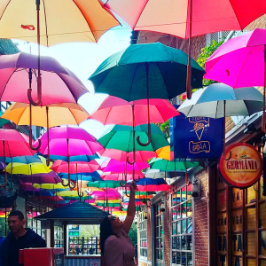 Foto e uma rua à frente do Shopping com alguns guarda-chuvas pendurados no "teto" bem coloridos e com uma menina posando para a foto segurando um deles