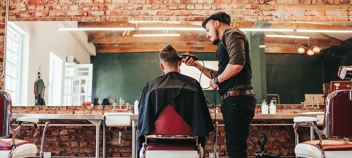 Imagem principal do post. Homem de costa recebendo um corte de cabelo em barbearia despojada, paredes de tijolo.