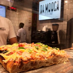 pizza em balcão com massa grossa e ao fundo funcionários e uma placa grande luminosa com o nome da pizzaria